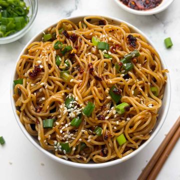 Chili oil garlic noodles served in a white bowl and green onions, sesame seeds and crispy chili oil served in small bowl in the side along with a pair of chop sticks.