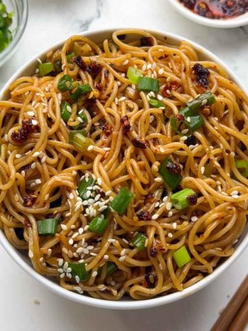 Chili oil garlic noodles served in a white bowl and green onions, sesame seeds and crispy chili oil served in small bowl in the side along with a pair of chop sticks.