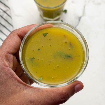 A person holding drumstick soup served in a glass container and another soup glass on the table.