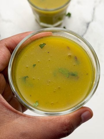 A person holding drumstick soup served in a glass container and another soup glass on the table.