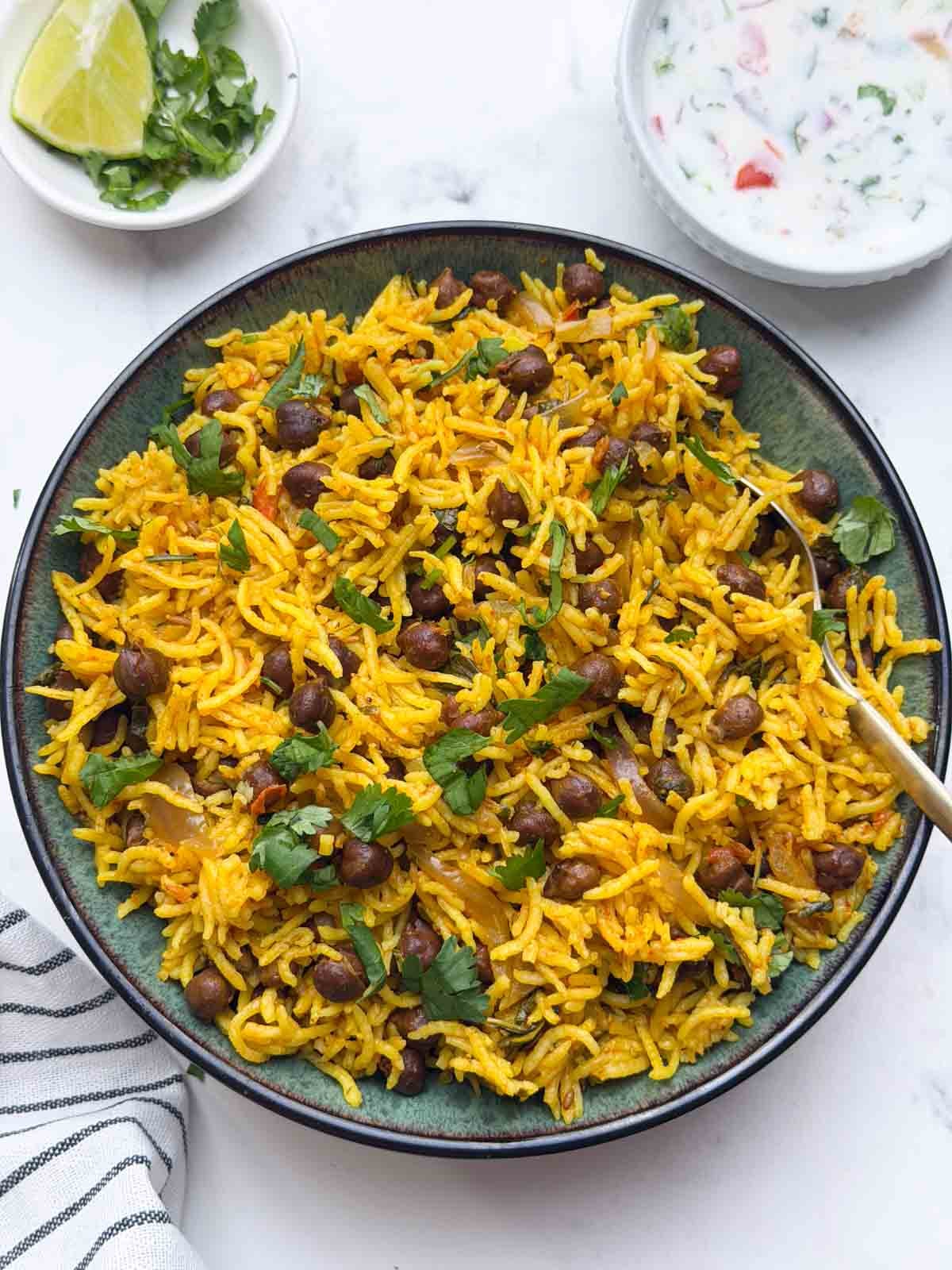 kala chana pulao served in a bowl with a spoon, raita and small bowl with lemon wedge and coriander leaves.