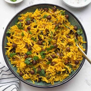 Black chana pulao served in a bowl with a spoon, raita and small bowl with lemon wedge and coriander leaves.