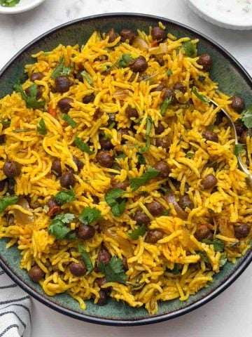 Black chana pulao served in a bowl with a spoon, raita and small bowl with lemon wedge and coriander leaves.