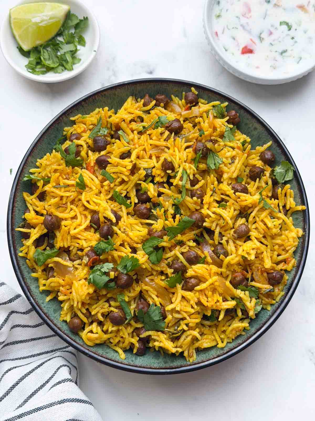 kala chana pulao served in a bowl with raita and small bowl with lemon wedge and coriander leaves.