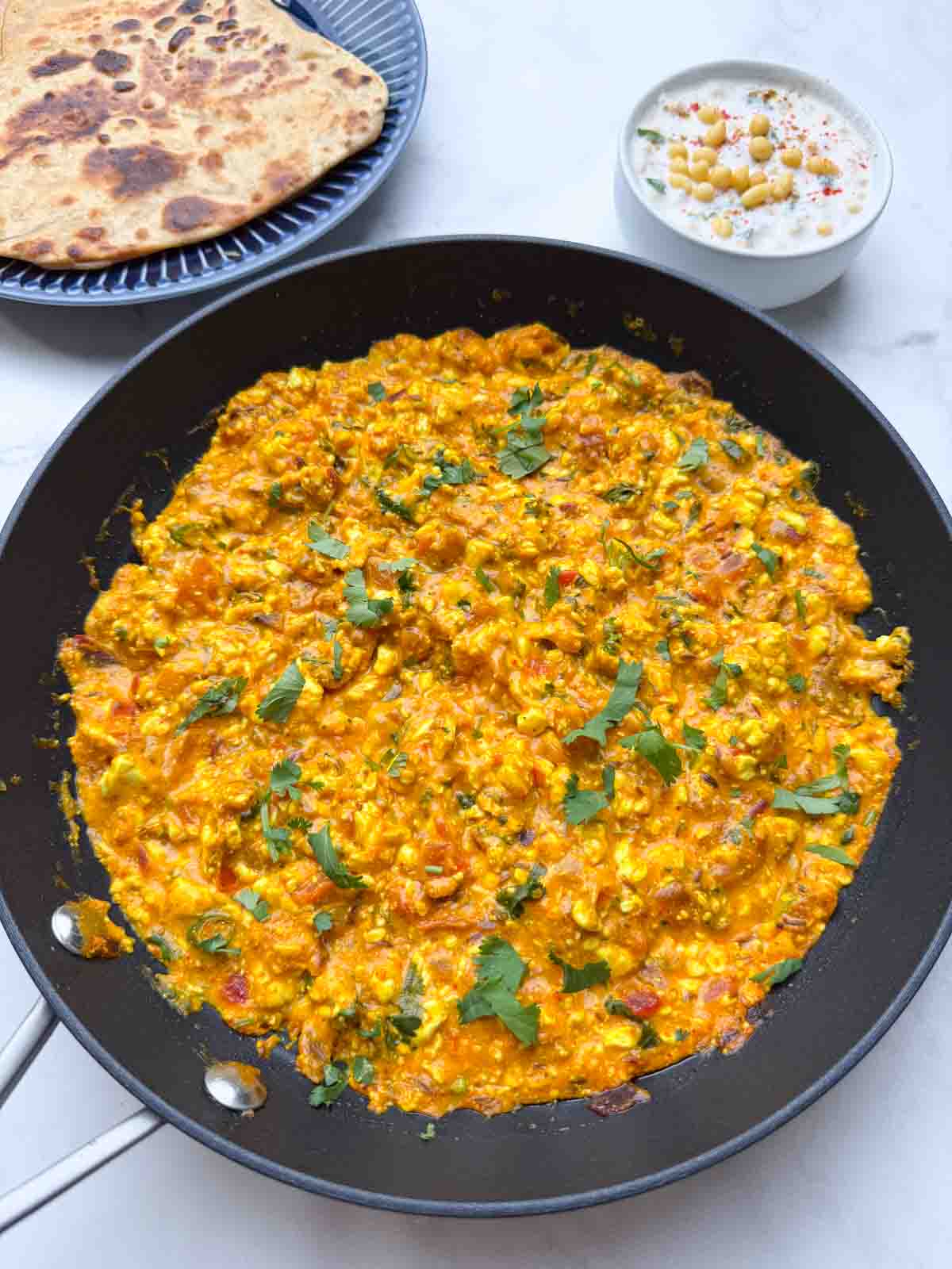 cottage cheese bhurji curry served in a pan with raita and paratha on the side.