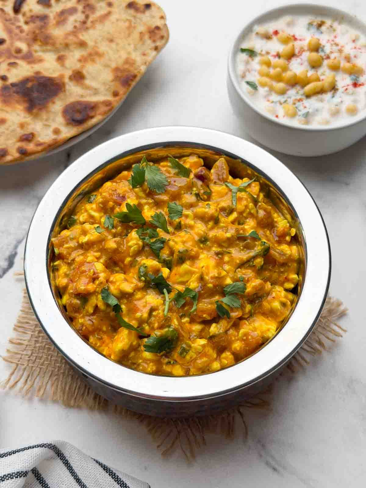 Cottage cheese curry served in a steel serving bowl with boondi raita and paratha on the side.