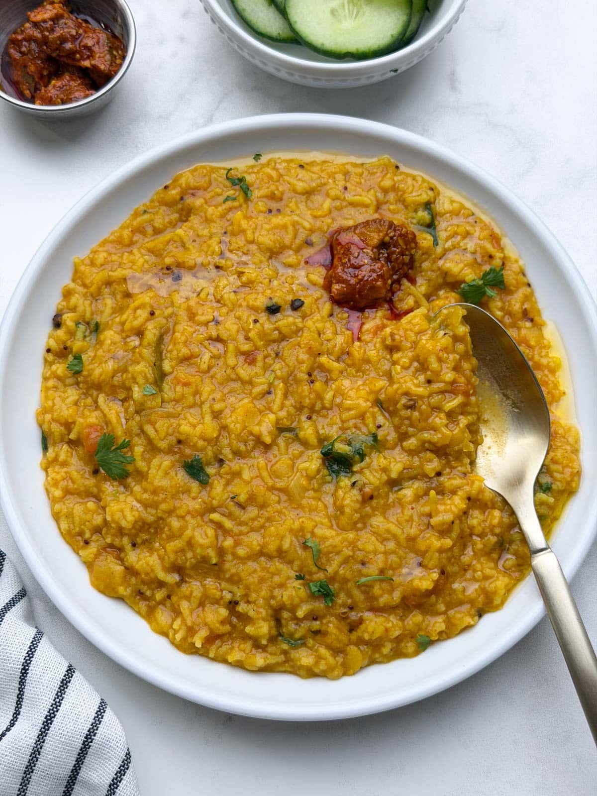 One pot rasam rice served in a white plate with pickle and spoon on it and cucumber and pickle on the side.