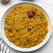 One pot rasam rice served in a white plate with pickle on it and cucumber and pickle on the side.