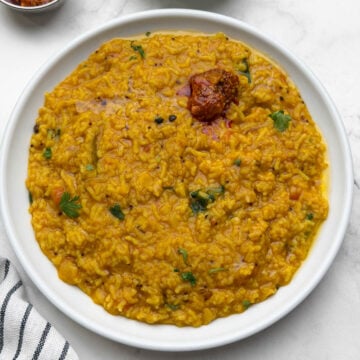 One pot rasam rice served in a white plate with pickle on it and cucumber and pickle on the side.
