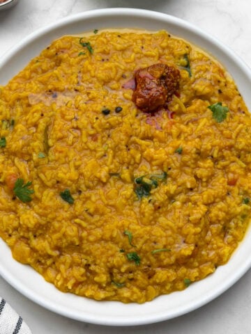 One pot rasam rice served in a white plate with pickle on it and cucumber and pickle on the side.