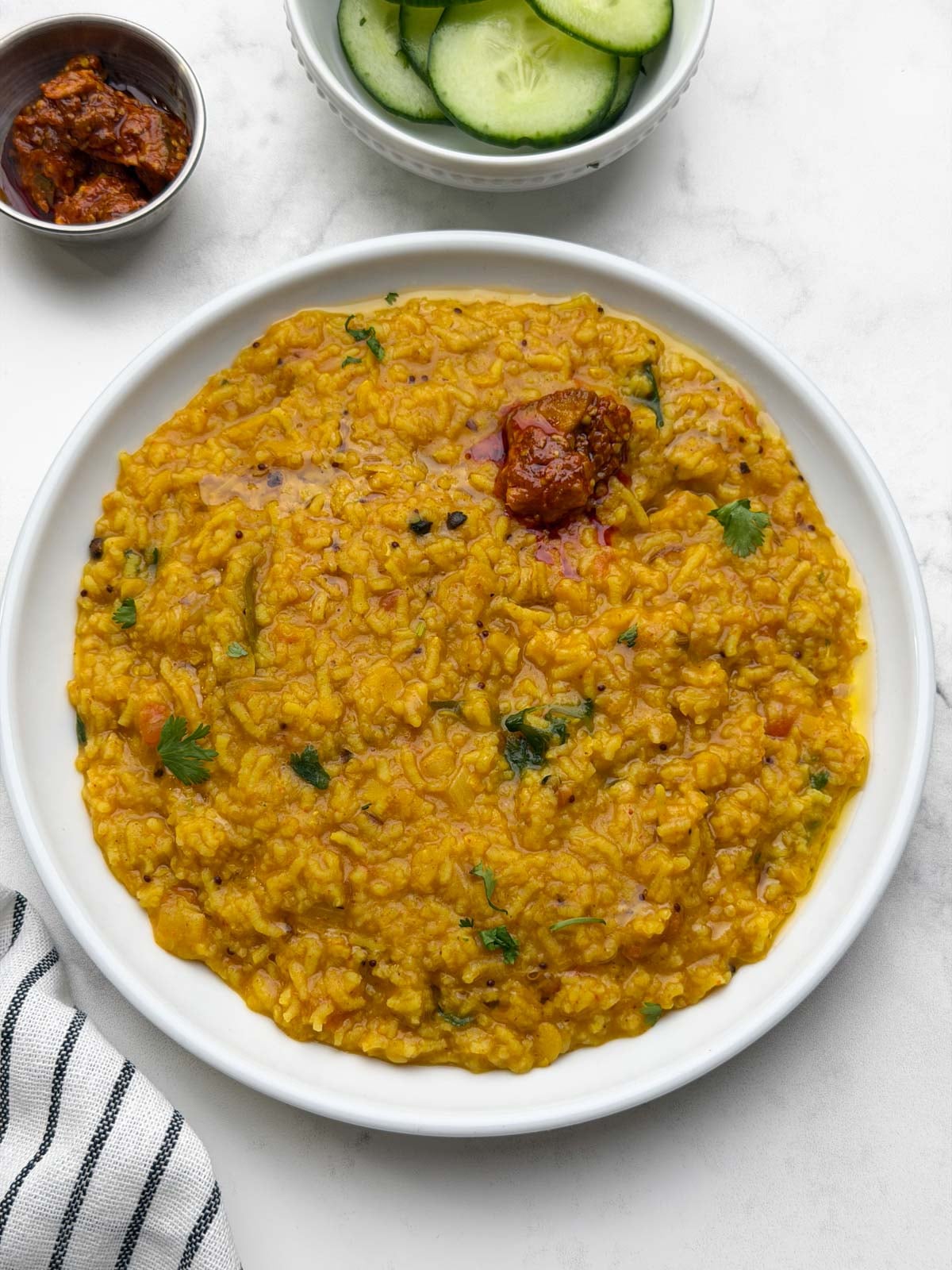 One pot rasam rice served in a white plate with pickle on it and cucumber and pickle on the side.