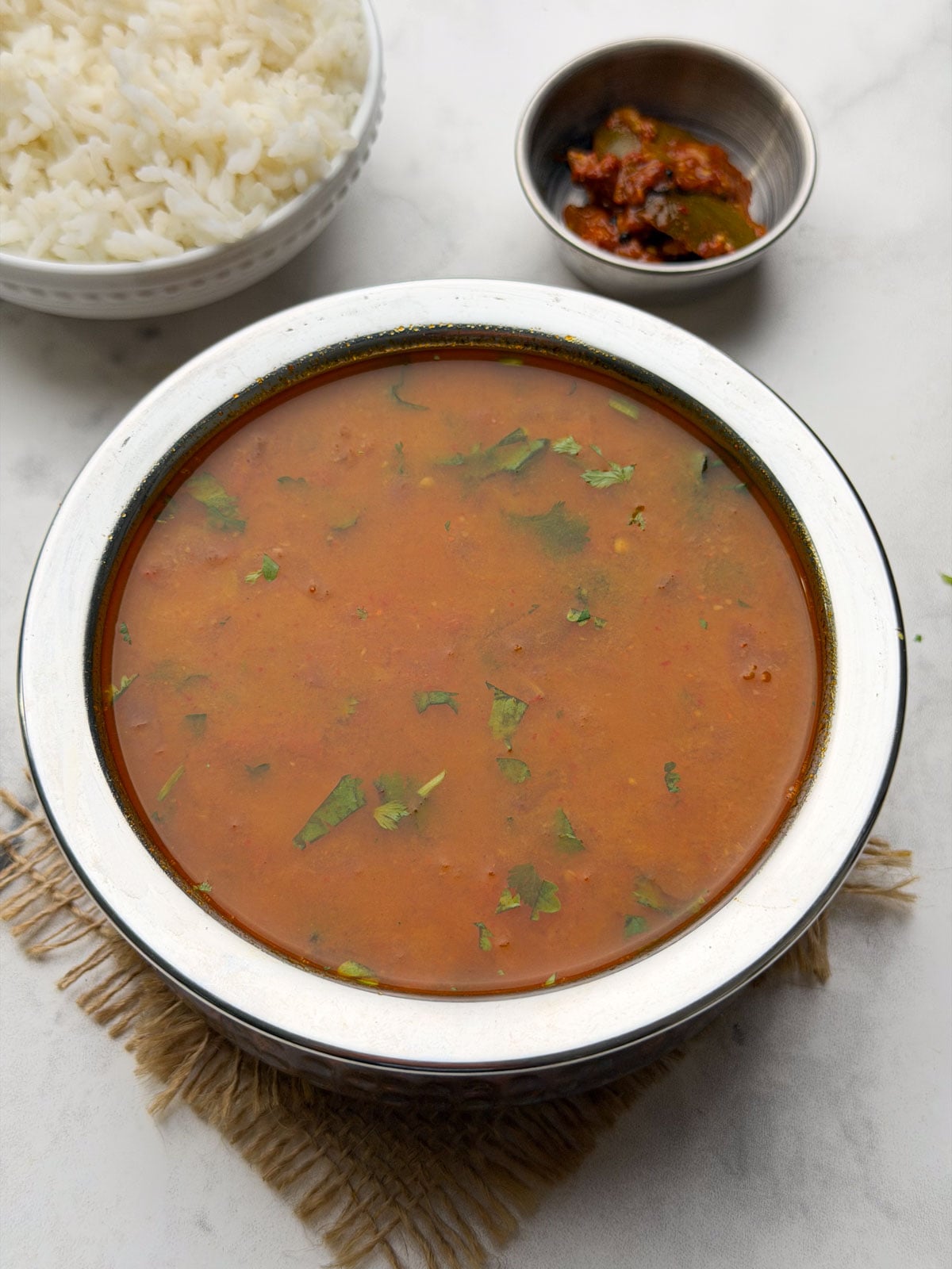 Tomato saaru served in a steel bowl with a bowl of steamed rice and pickle on the side.