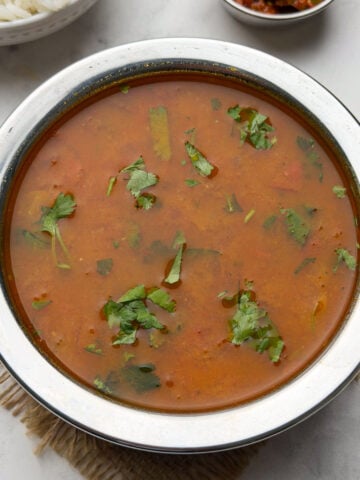Tomato saaru served in a steel bowl with a bowl of steamed rice and pickle on the side.